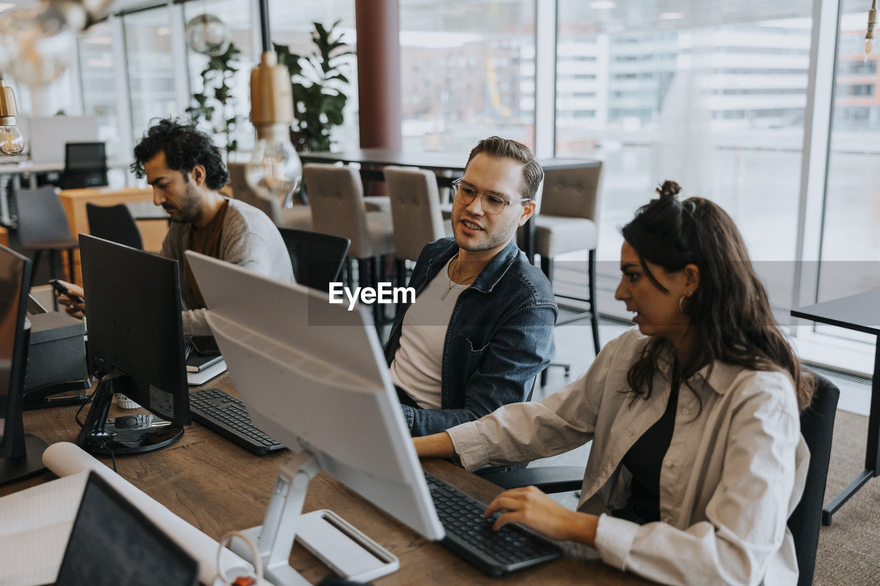 Young male and female colleagues discussing over computer while sitting by businessman at desk in office