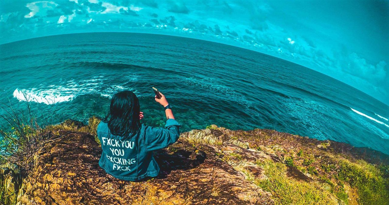 Rear view of woman photographing sea while sitting on cliff