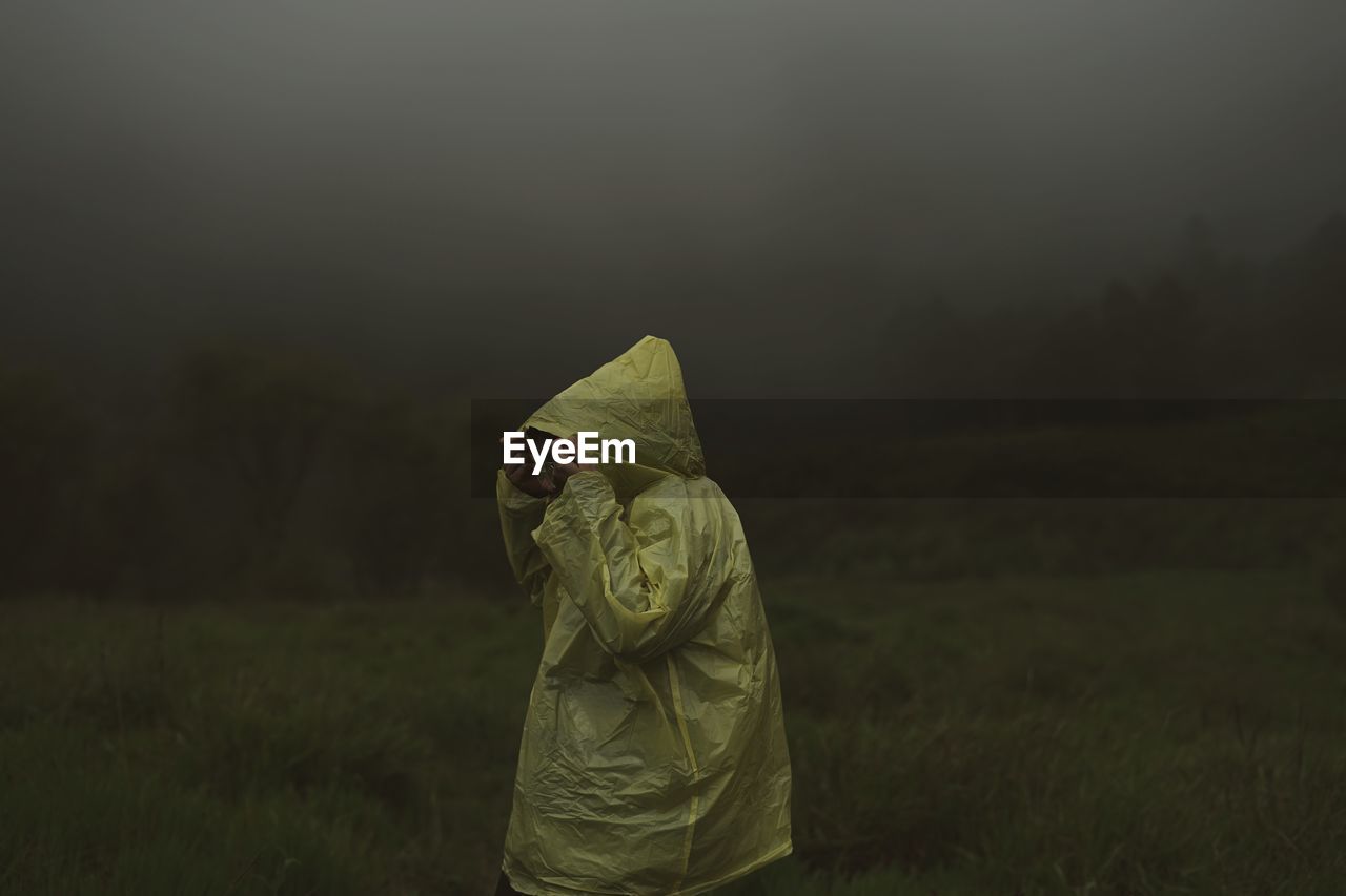 A woman standing and wearing yellow plastic jacket on field against misty cloud during rainy season