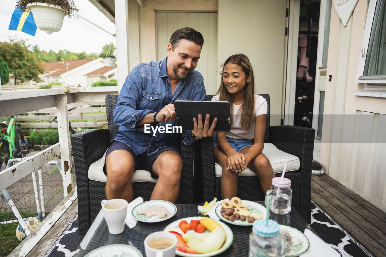 Father sharing tablet pc with daughter sitting on chair at porch