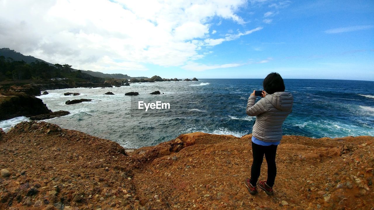 Woman photographing sea while standing on rock formation against sky