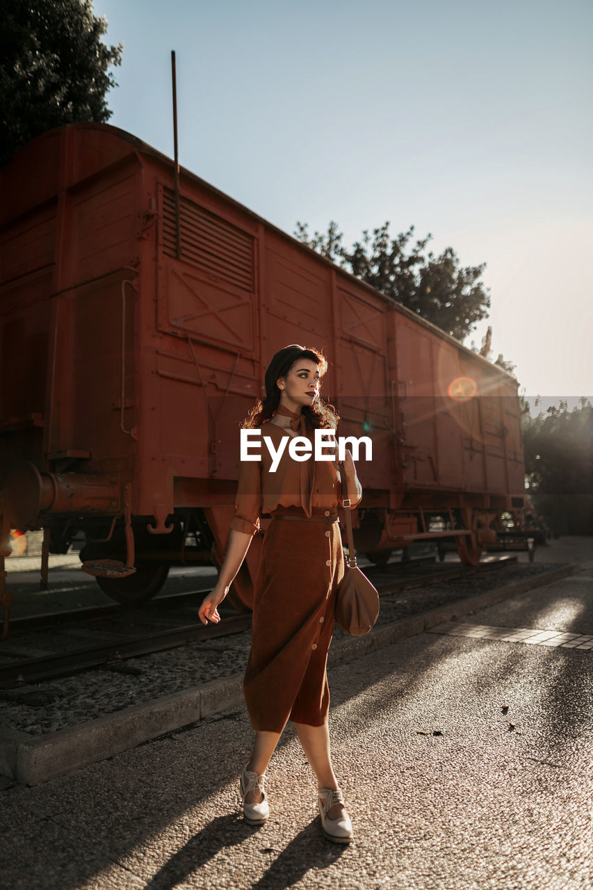 Dark haired female totally wearing terracotta clothes in vintage style standing near terracotta car train and holding open book in hands having dreaming look