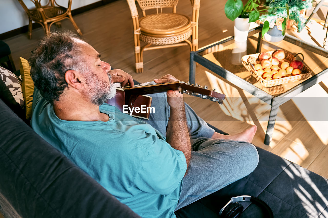 Middle-aged bearded man playing acoustic guitar while sitting on sofa in light living room.