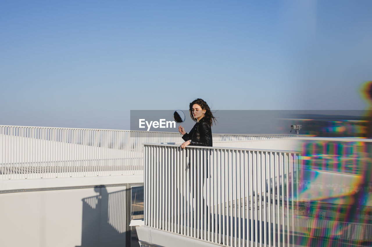 Side view of young woman holding mirror looking away while standing on terrace against clear blue sky