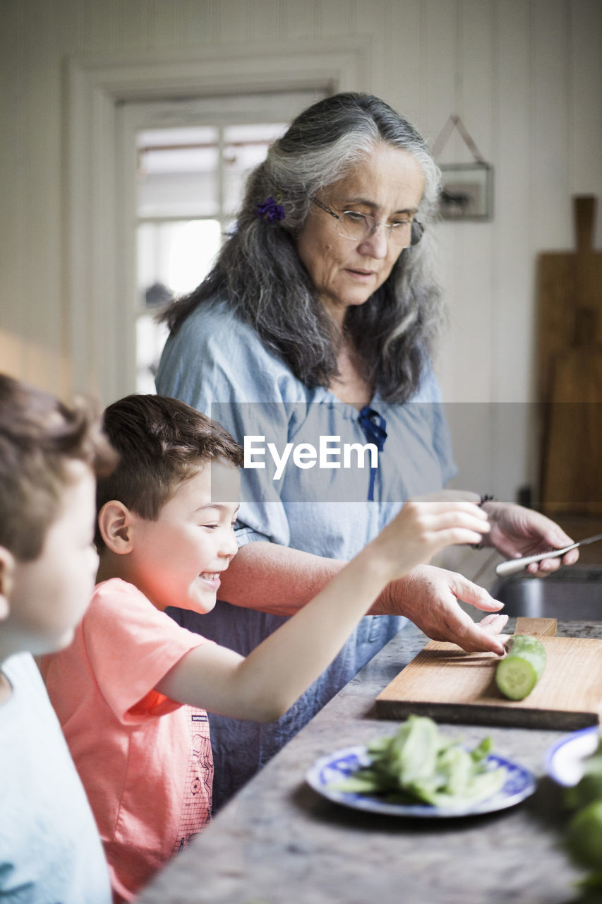 Grandmother and grandsons cutting cucumber in kitchen at home