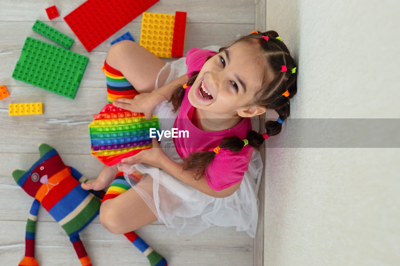 Cheerful little girl in bright colorful clothes, sits at home, on the floor