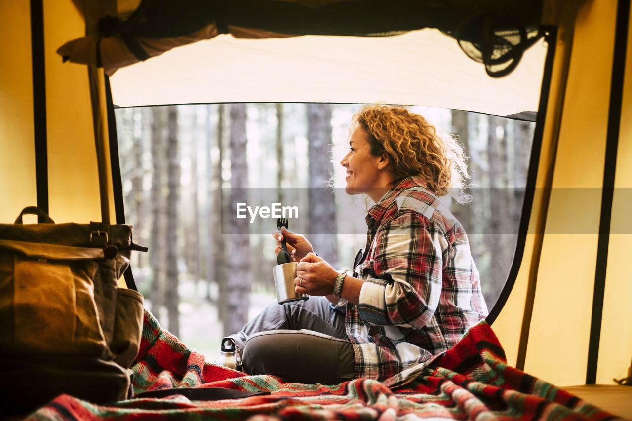 Side view of smiling woman holding mug while sitting by tent in forest