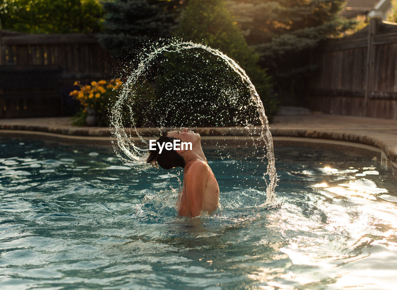 Teenage boy flipping water from his hair in an arc in a backyard pool.