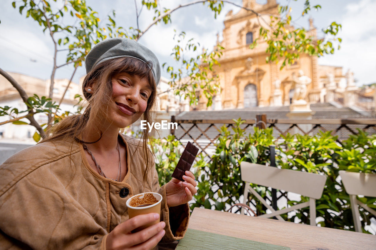 portrait of young woman using mobile phone while sitting on table against building