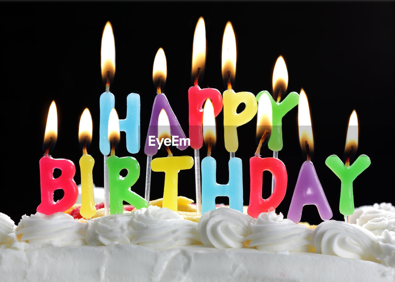 Close-up of illuminated happy birthday candles on cake against black background