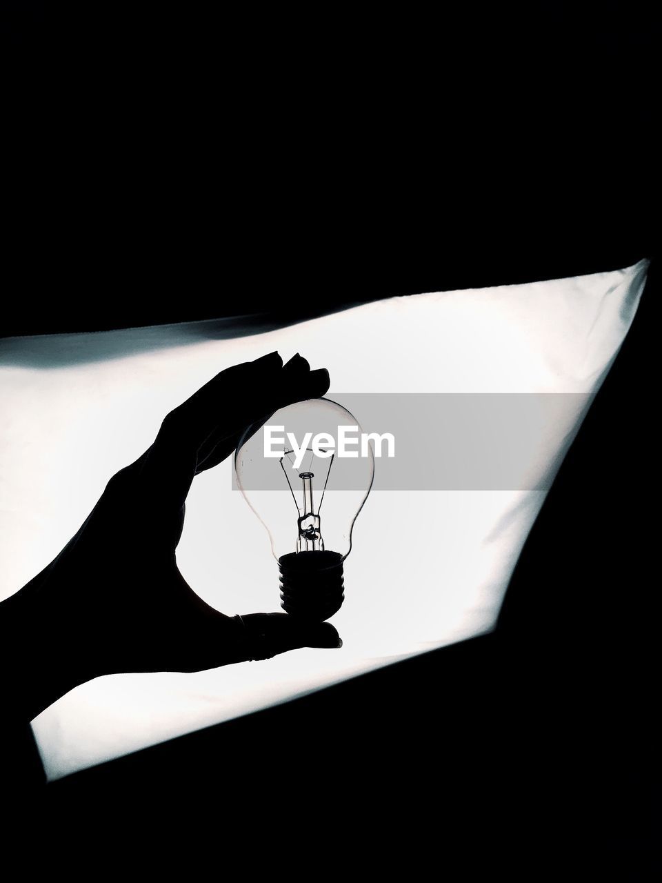 Silhouette cropped hand holding light bulb against illuminated light