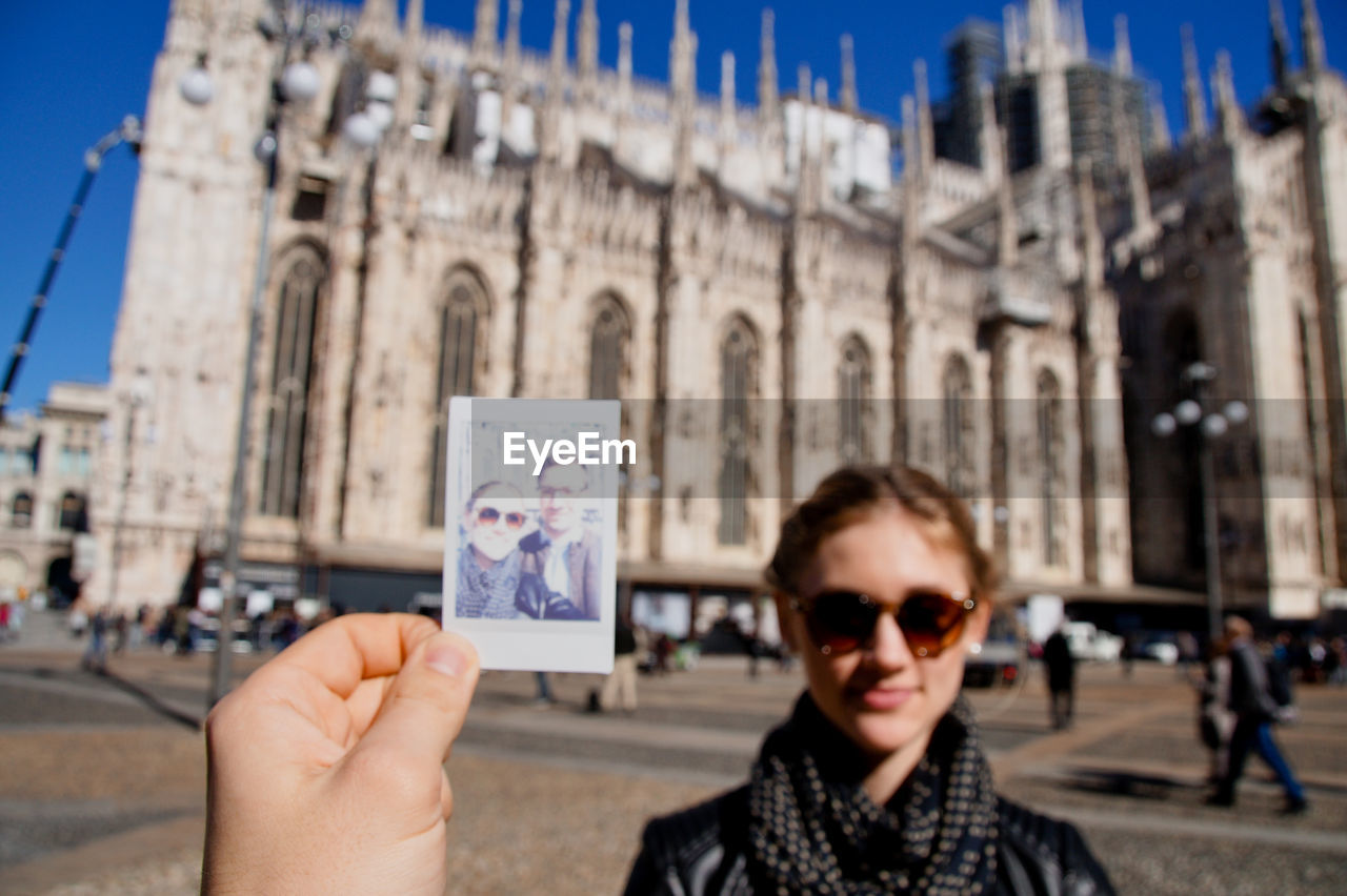 Cropped hand of man holding photograph with woman in background against milan cathedral