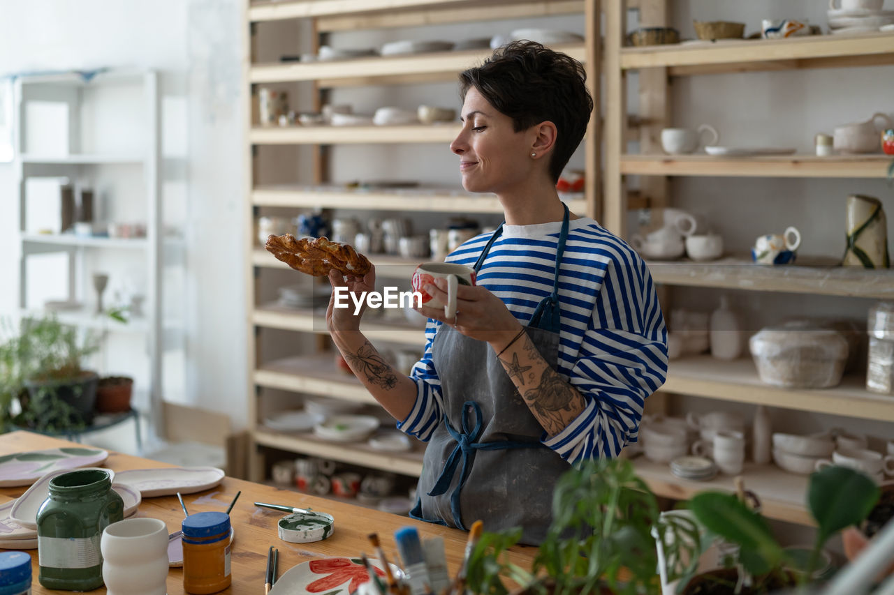 Artisan tattooed girl drink coffee and looking at tasty bun lustily in art studio during break