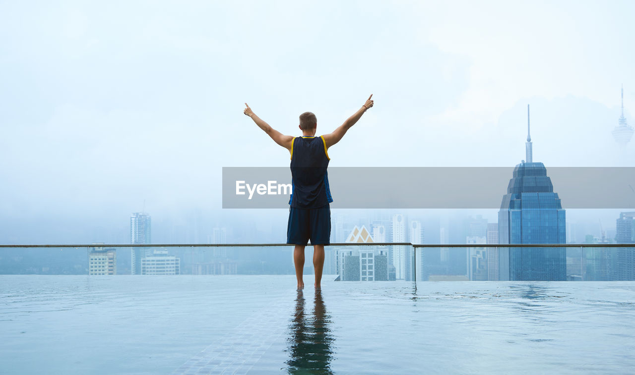 Man standing in infinity pool against sky in city