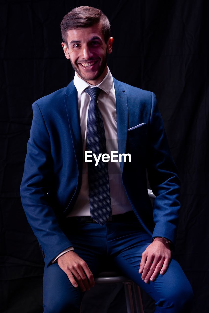 Portrait of smiling young businessman sitting over black background