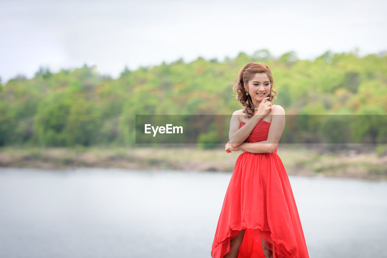 Smiling woman in red dress standing against lake