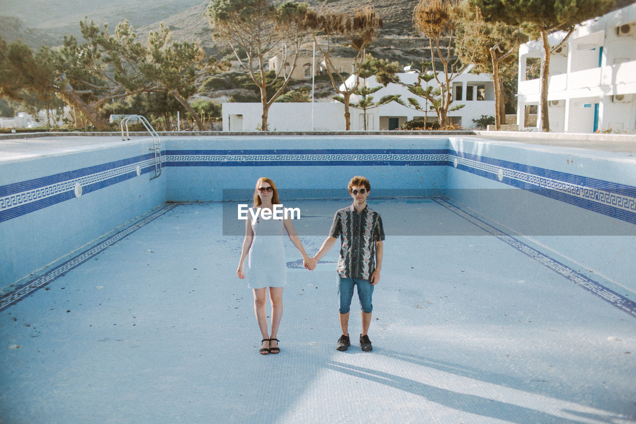 Full body young calm couple in casual outfits holding hands and looking at camera while standing in pool without water in sunny resort
