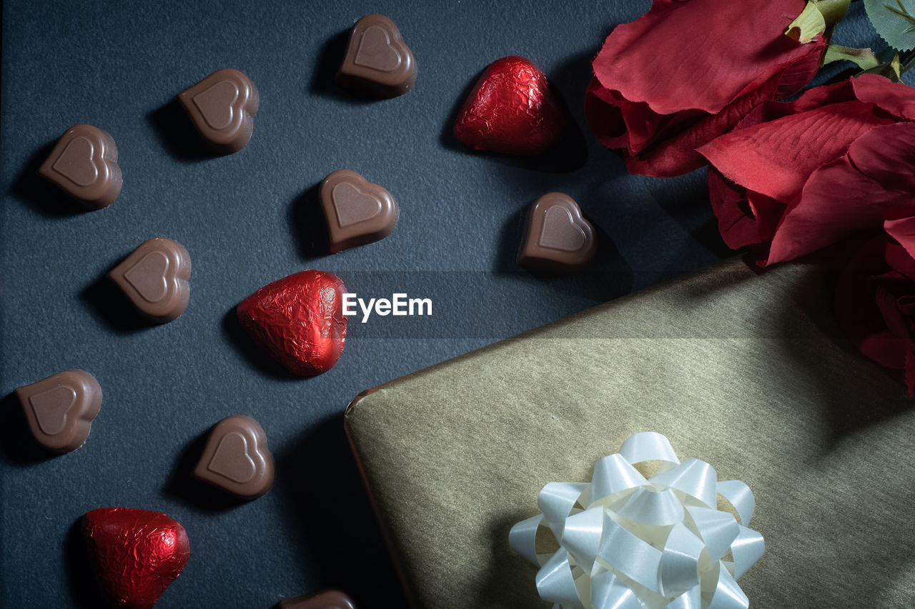 Closeup of heart-shaped chocolate confections, red roses, and a gift against a dark background.