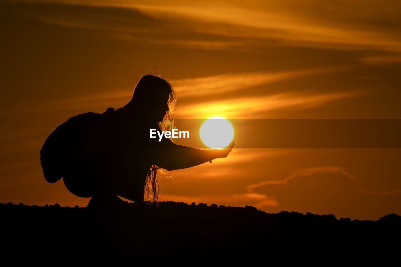 Optical illusion of silhouette woman holding sun while standing against orange sky during sunset