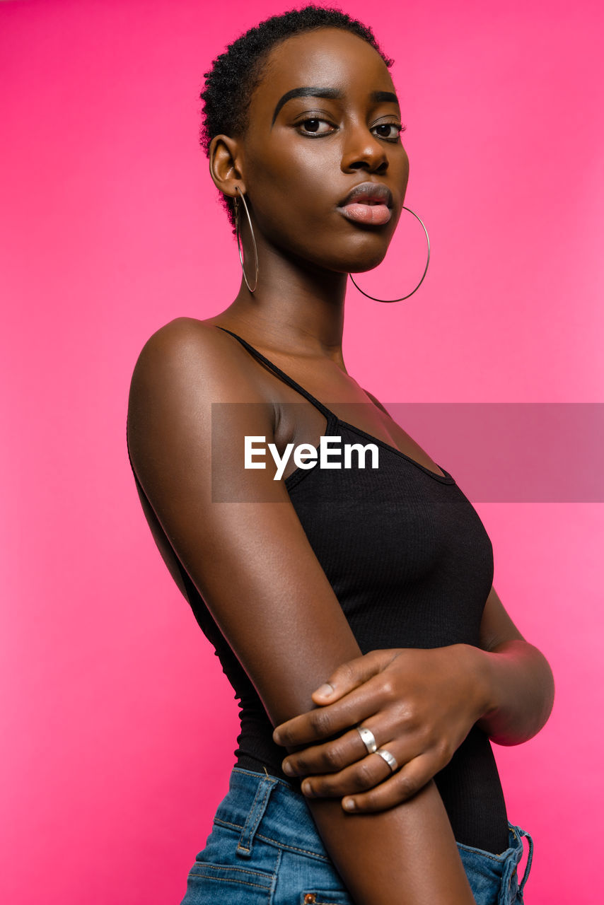 Confident young fit african american female model in black top and jeans looking at camera while standing against pink background