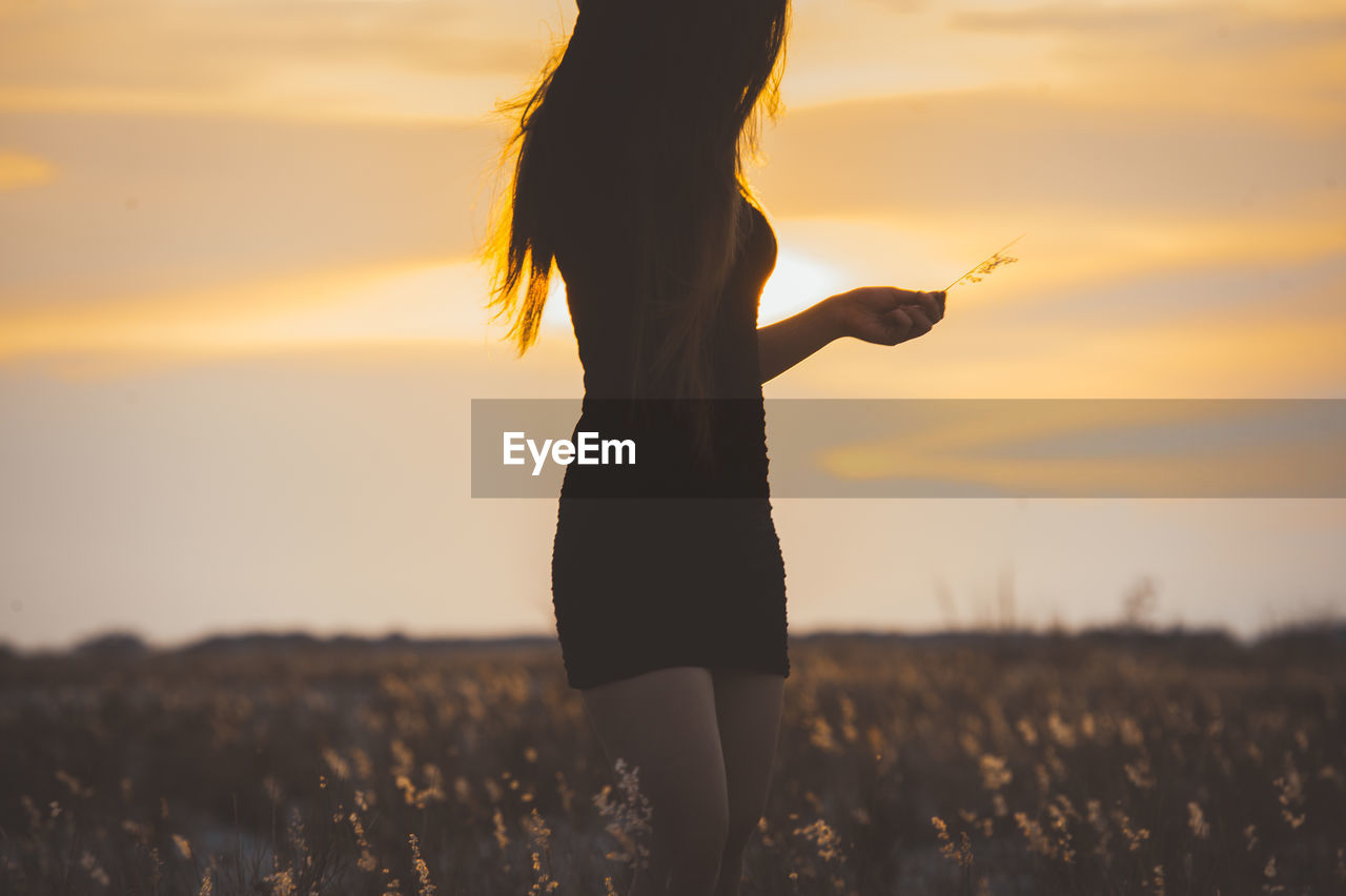 Midsection of woman standing amidst field against sky during sunset