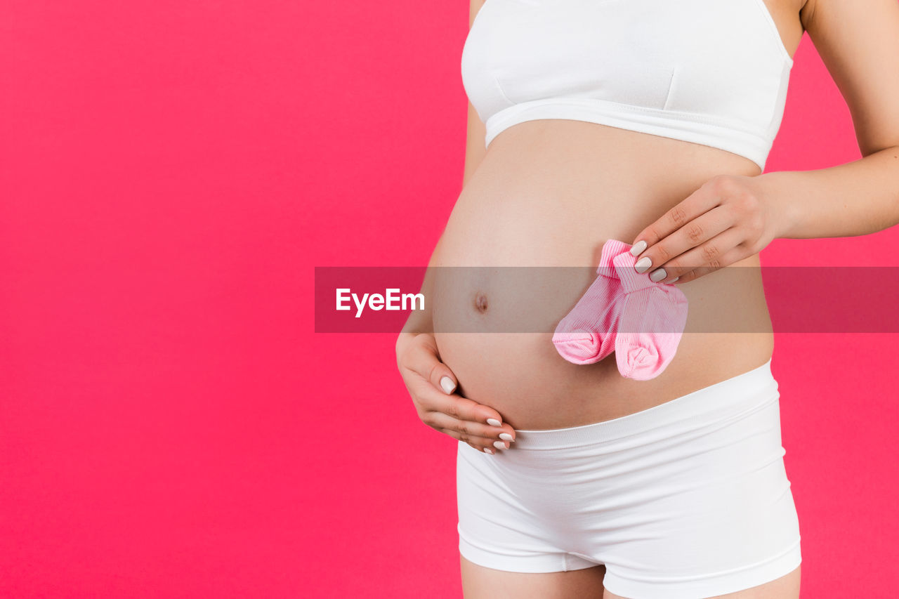 Pregnant woman in underwear holding pink socks for a baby girl at background. childbirth expecting.