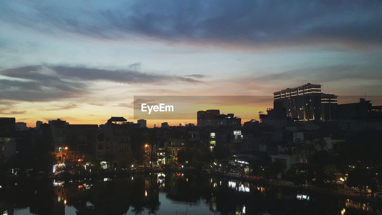 Illuminated buildings by river against sky at sunset