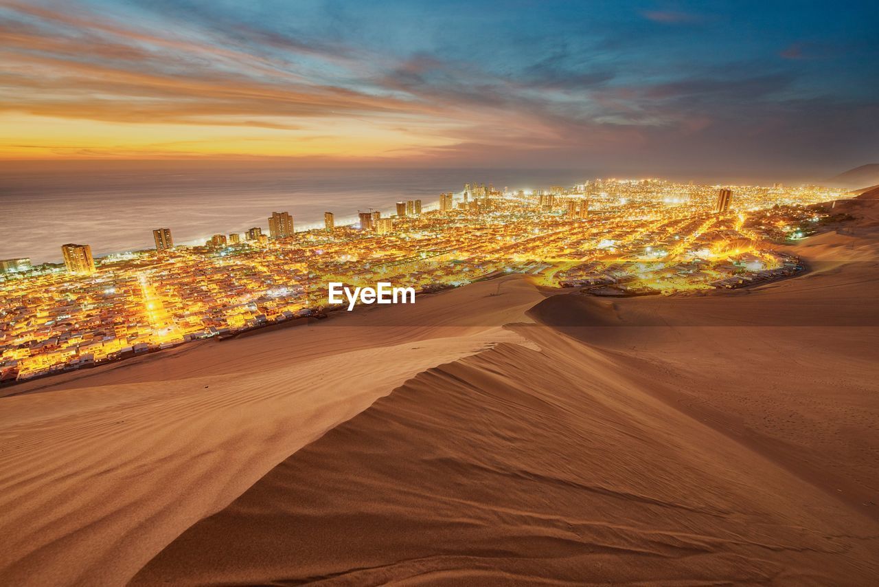 Desert by illuminated cityscape and sea against sky during sunset