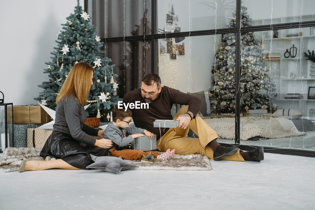 Family with child playing near christmas tree. child unpacking gifts, parents enjoy christmas tree