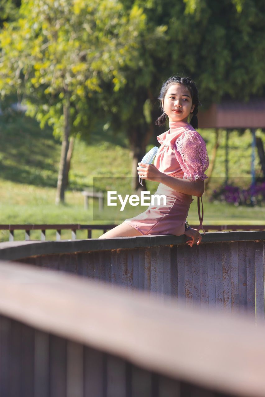 Portrait of a smiling young woman sitting outdoors and holding face mask on hand.