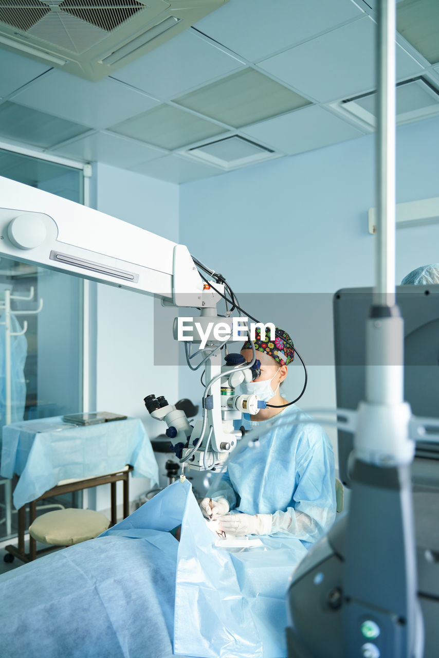 Attentive female doctor in surgical uniform and sterile mask looking through microscope while operating eye of unrecognizable patient in hospital