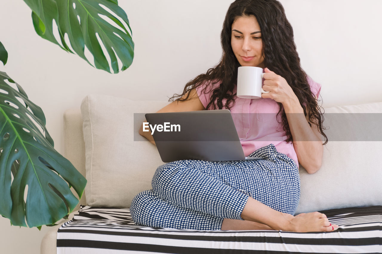 Woman with dark curly hair surfing the net while sitting on the couch and drink coffee at home. 
