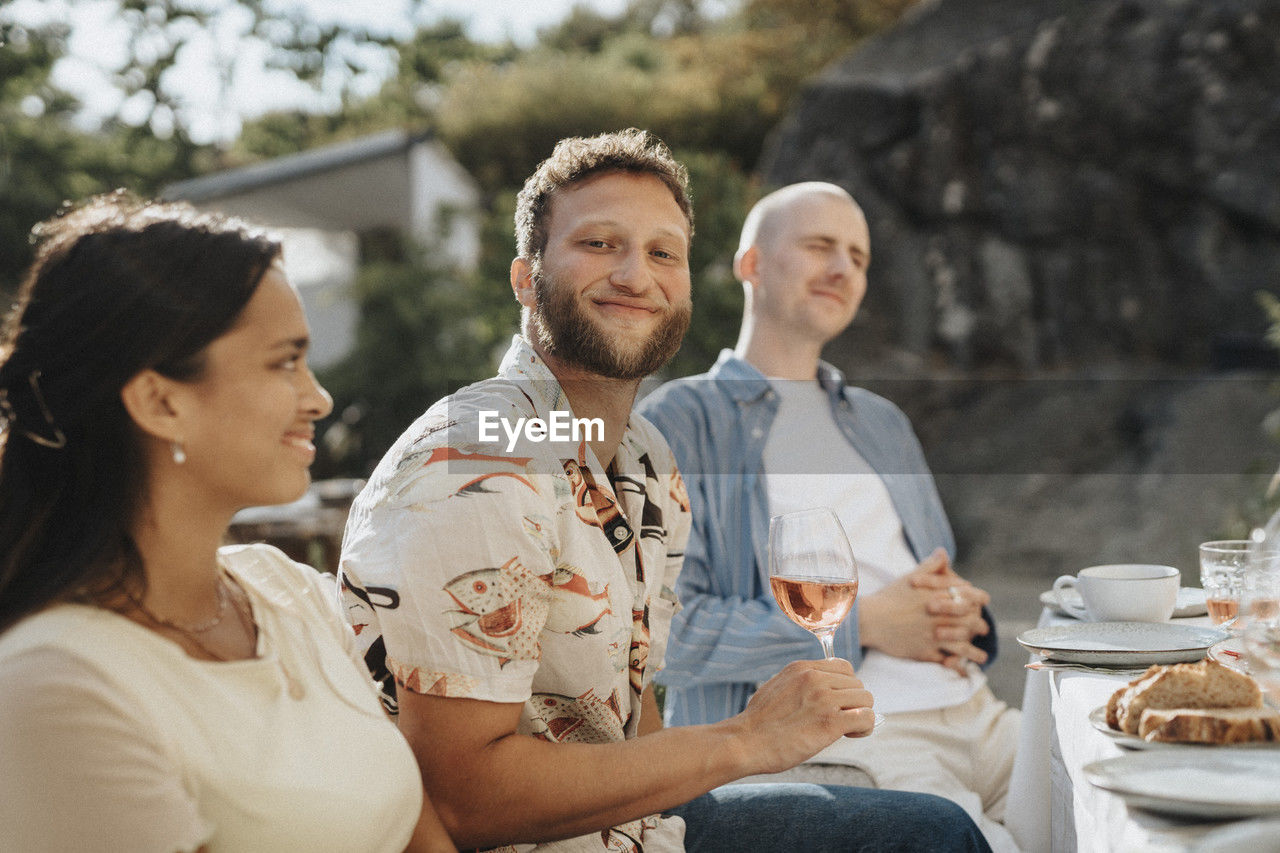 Portrait of smiling young man holding wineglass while sitting with friends during dinner party at cafe