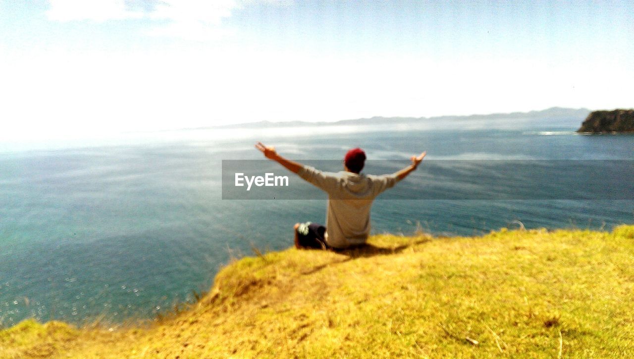 Rear view of man with arms outstretched sitting on cliff against sea