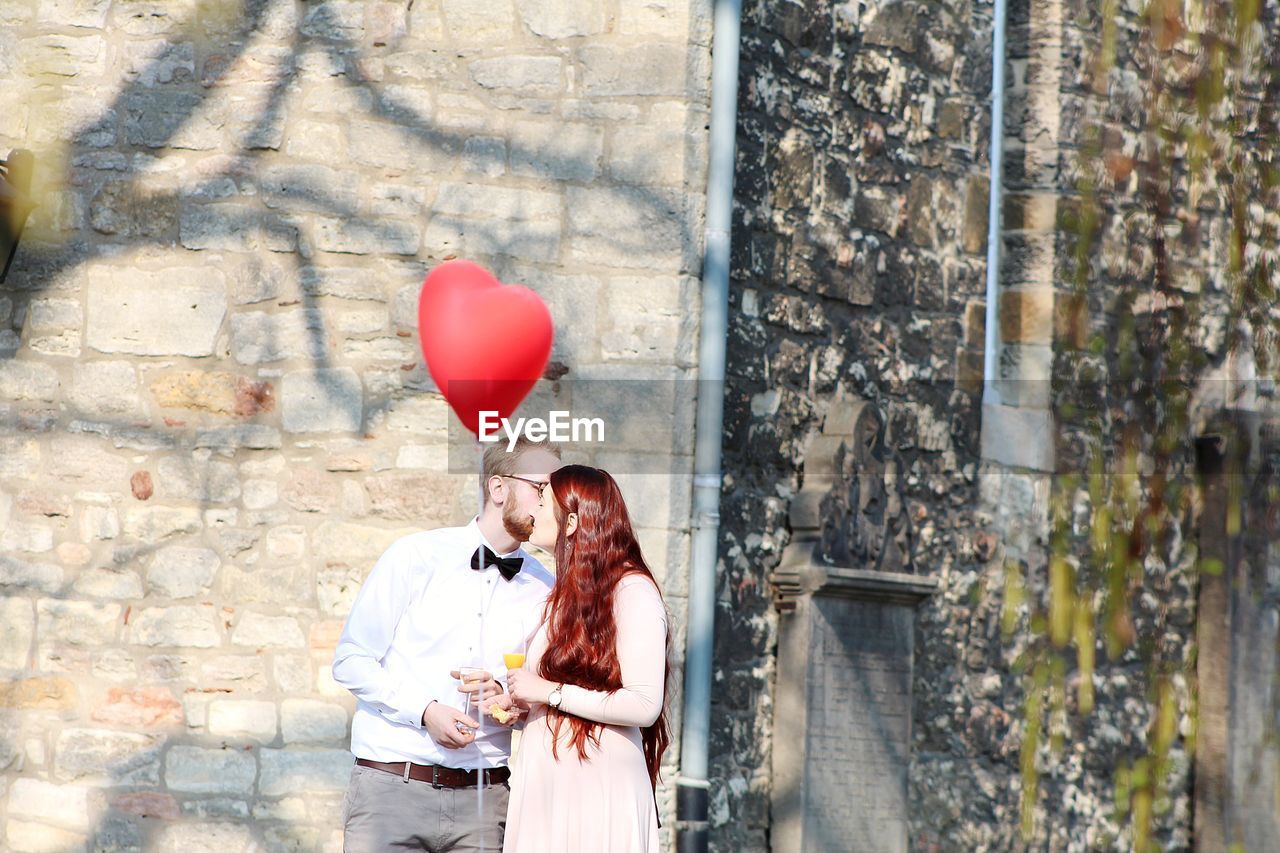 Heart shape balloon floating against couple kissing while holding drinks outdoors