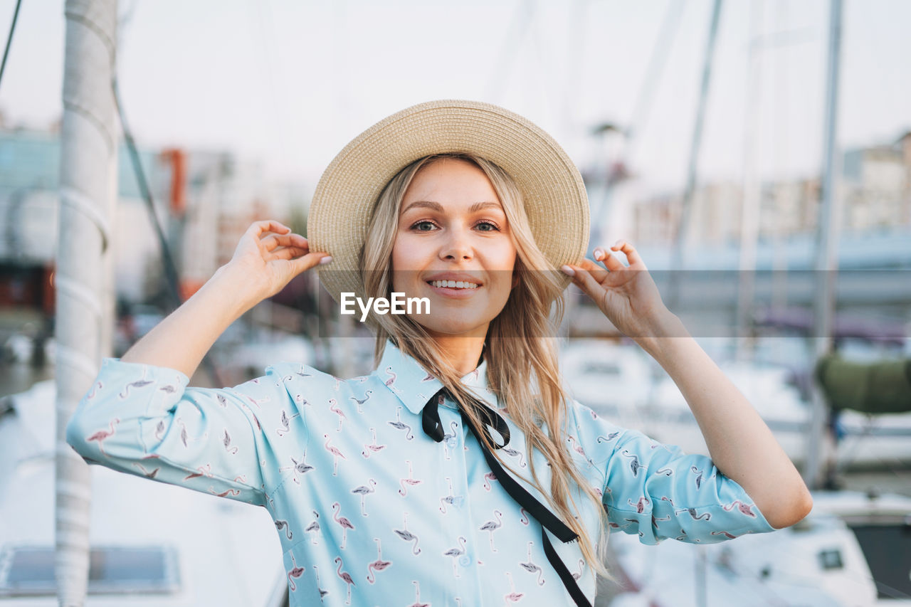 Beautiful smiling blonde young woman in blue dress and straw hat at pier in sunset time