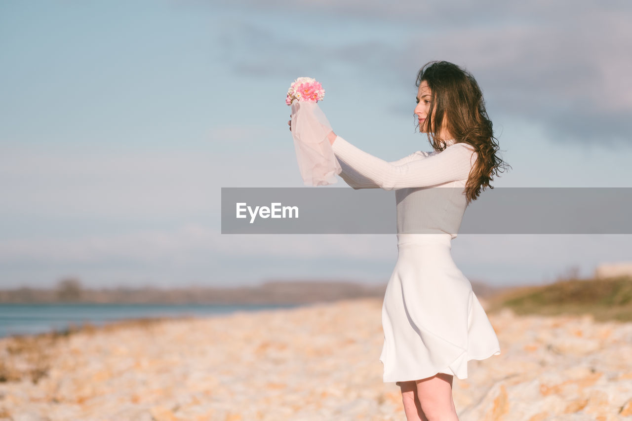 Portrait of young woman standing at beach against sky