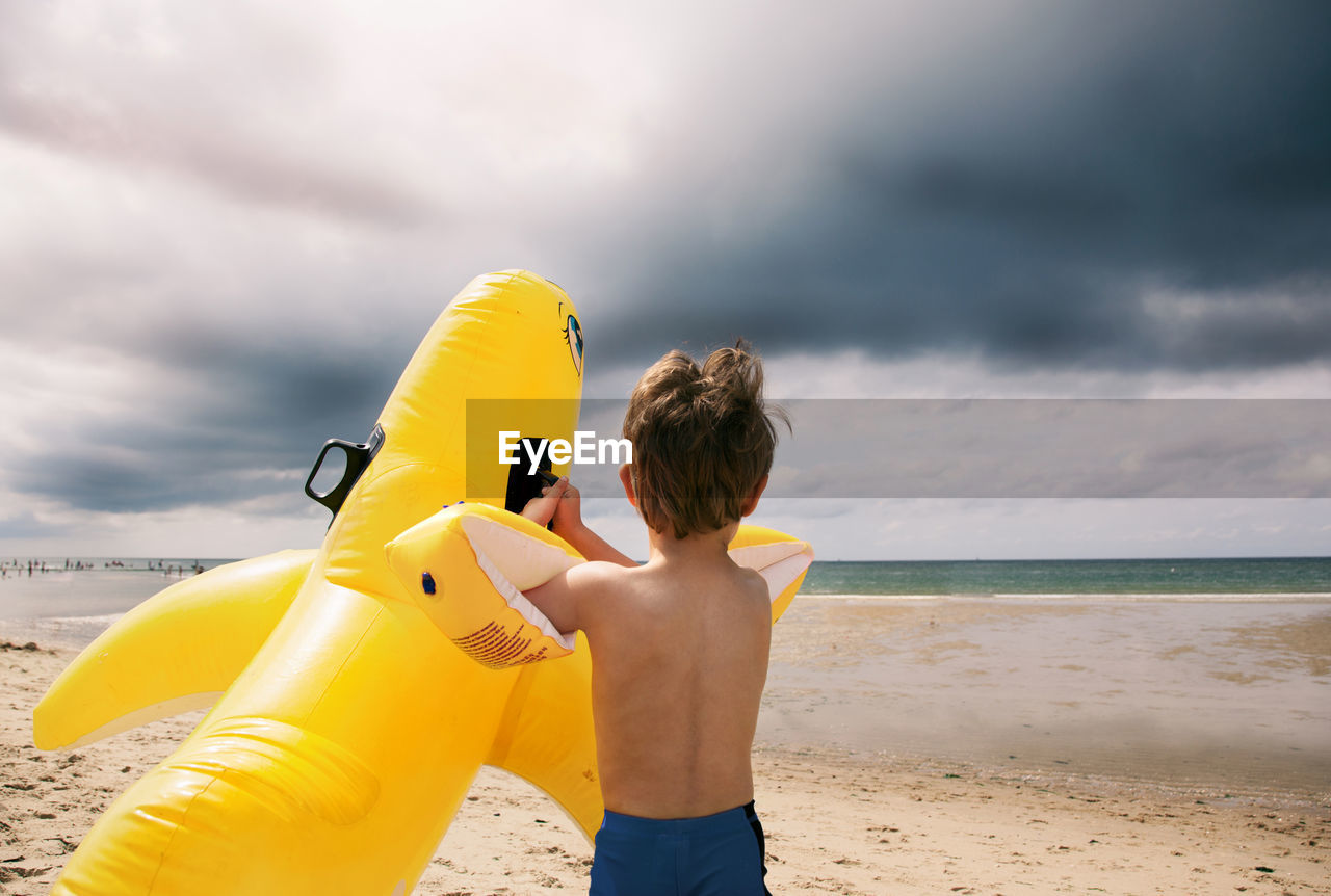 Rear view of shirtless boy playing at beach against cloudy sky