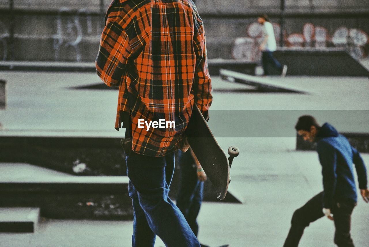 Man walking with skateboard in skateboard park with friends skating in background