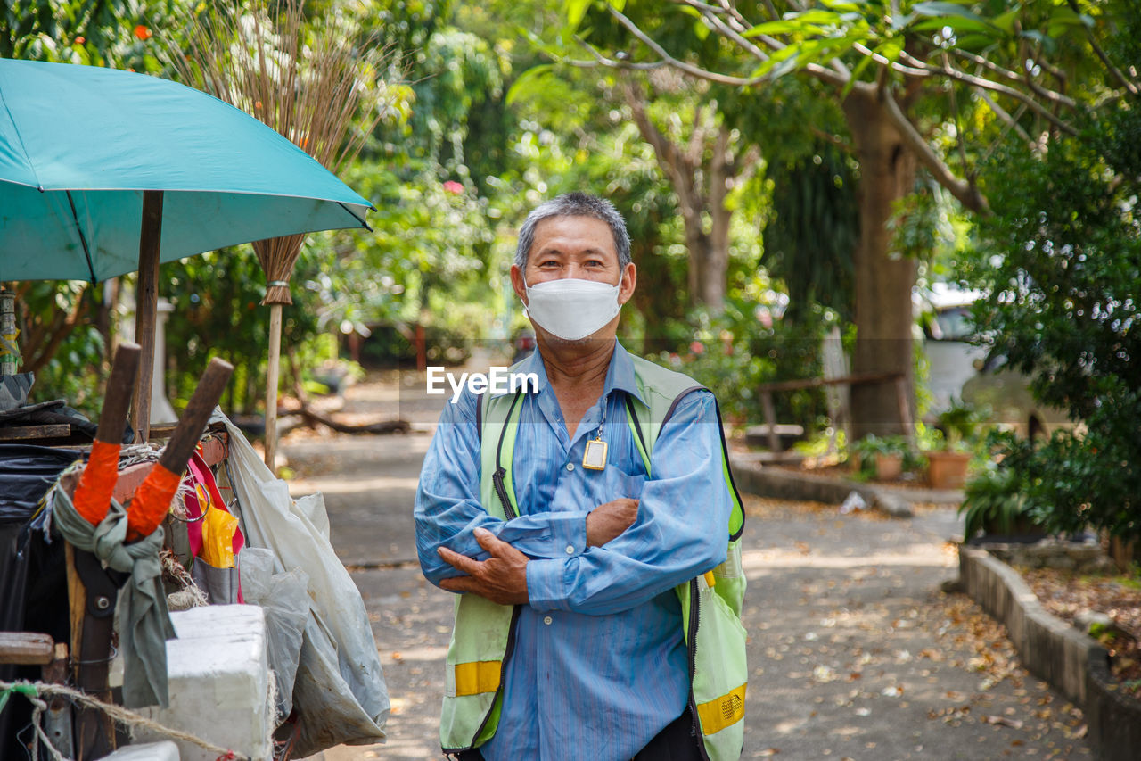 An asian man street cleaner wearing a mask standing with his arm crossed next to an old garbage cart
