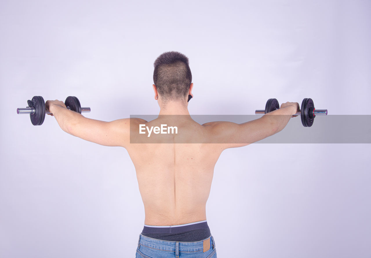 Rear view of shirtless man lifting dumbbells against purple background