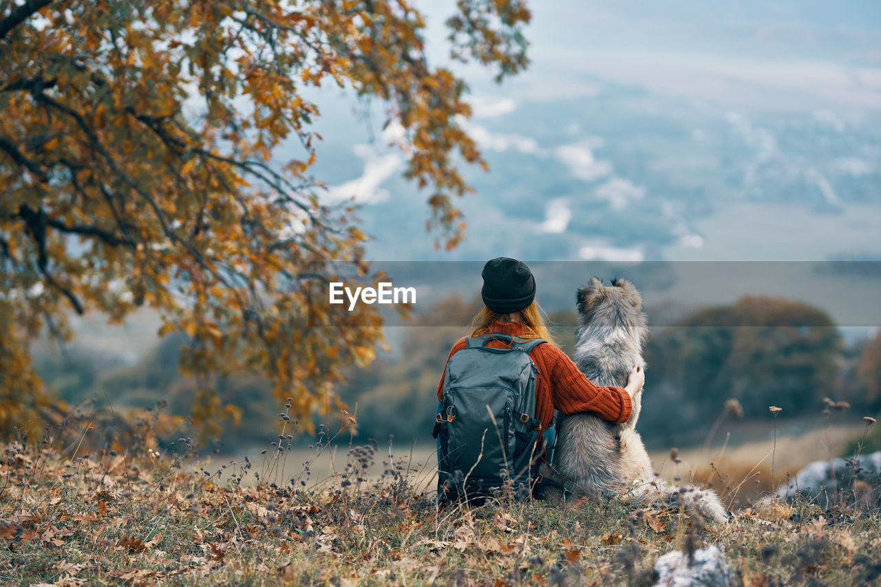 REAR VIEW OF MAN SITTING ON TREE DURING AUTUMN