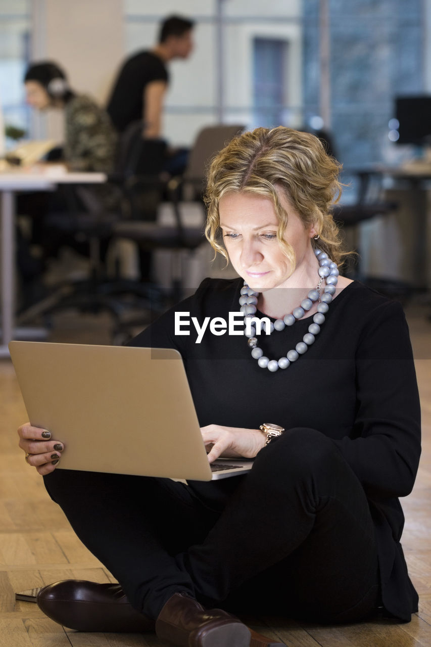 Full length of mature businesswoman using laptop while sitting on floor at office