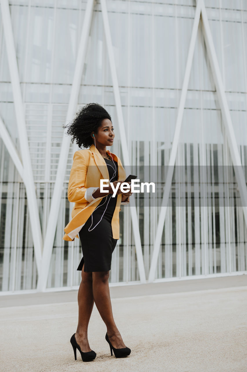 Female entrepreneur listening music through earphones while looking while standing against building