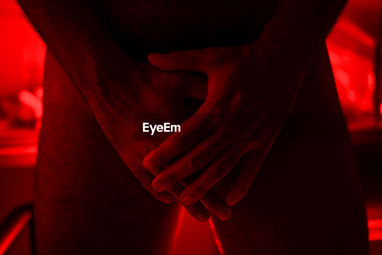 Midsection of naked man gesturing while standing in illuminated red room