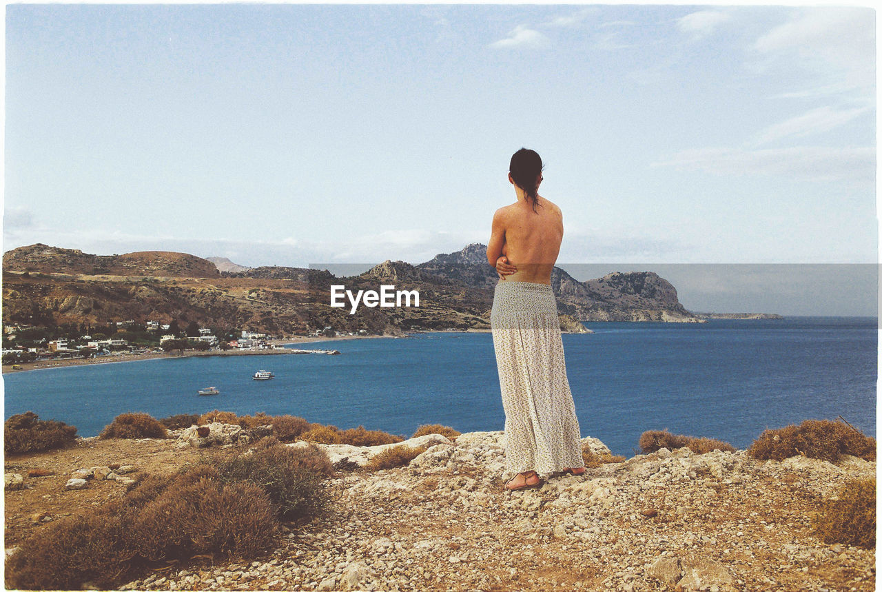 Rear view of shirtless woman standing on rock formation against sky