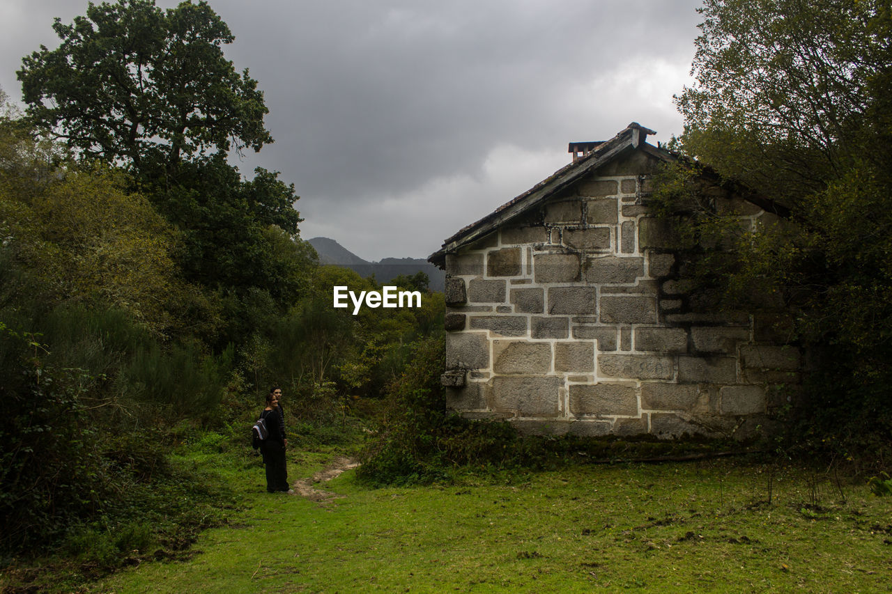 People standing on field by house and trees against sky