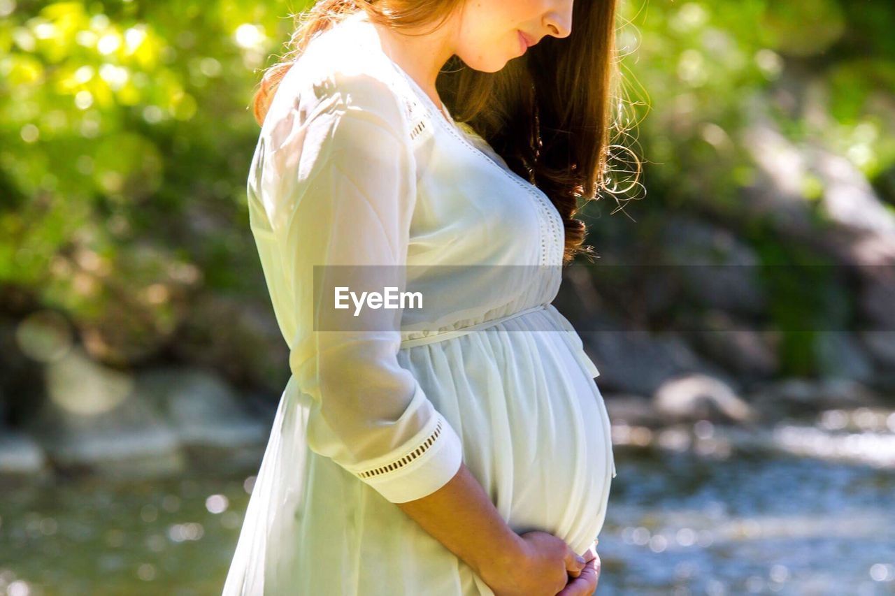 MIDSECTION OF WOMAN STANDING BY TREE AGAINST BLURRED BACKGROUND