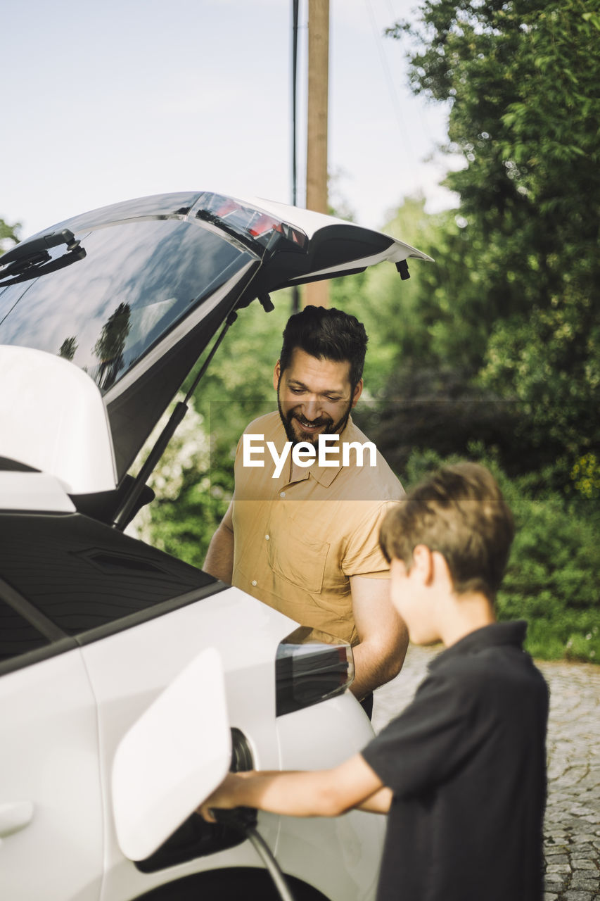 Smiling father standing by son charging electric car