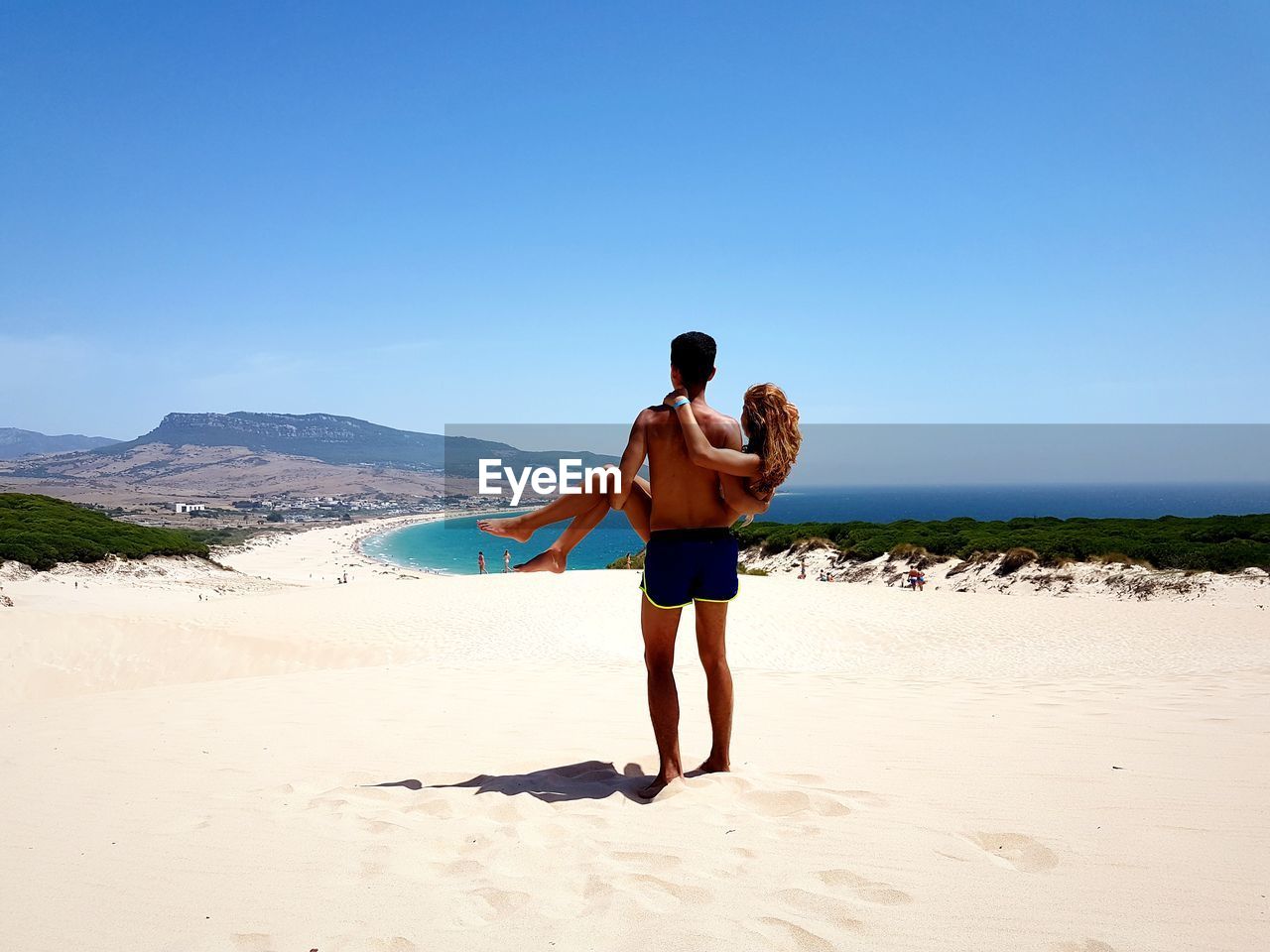 Rear view of shirtless man lifting girlfriend while standing at beach against clear sky